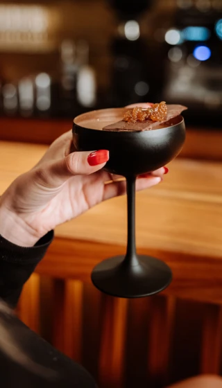 Elegantes Cocktailglas mit Schokoladenstück und kandierten Früchten, gehalten in einer Hand, vor einer stilvollen Bar.