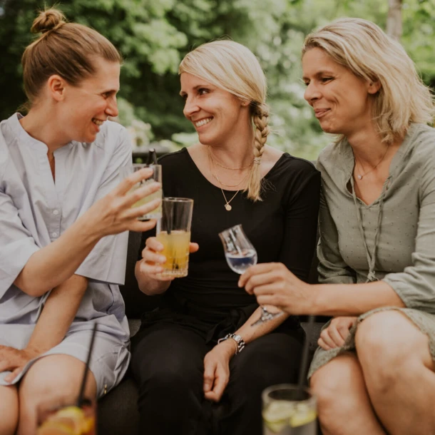 Three women smiling and toasting with drinks outdoors.