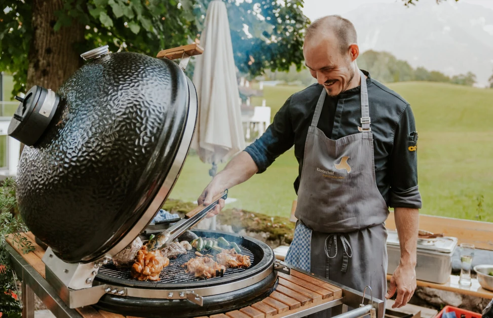 Ein Koch grillt Fleisch und Gemüse auf einem großen Grill im Freien, umgeben von einer schönen, grünen Landschaft.