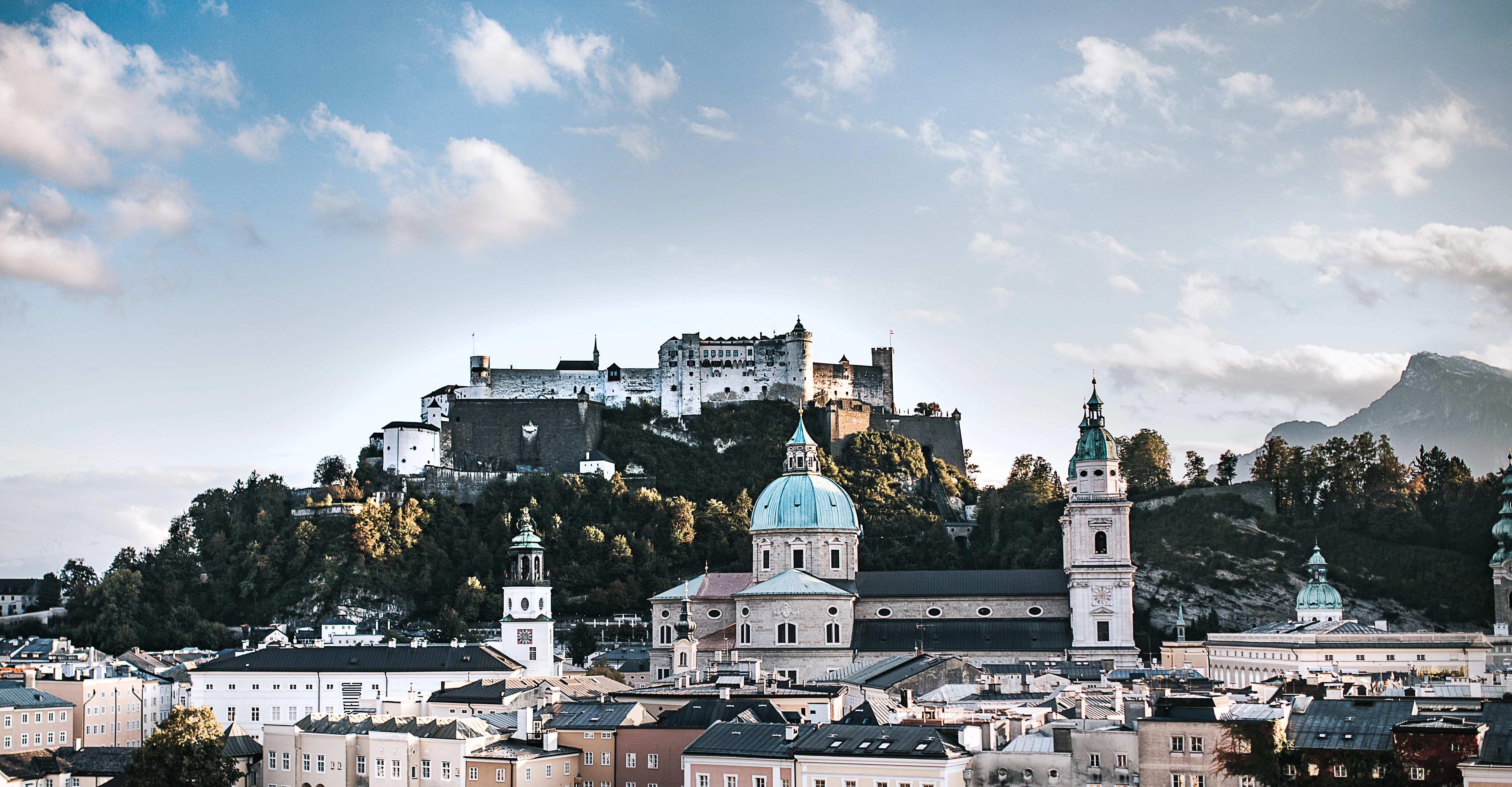 View of Salzburg with Hohensalzburg Fortress, surrounded by historic buildings and mountains under a blue sky.