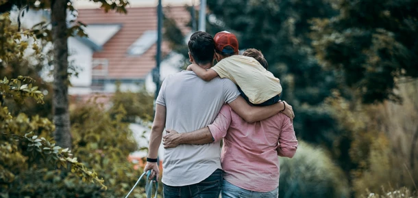 Ein Paar spaziert mit einem Kind auf dem Rücken durch eine grüne Allee, umgeben von Bäumen und Natur.