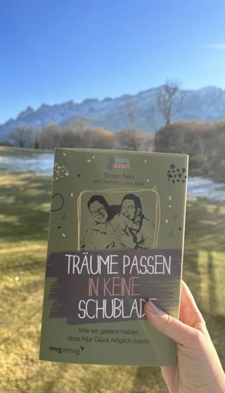Eine Person hält das Buch "Träume passen in keine Schublade" vor einer malerischen Berglandschaft.