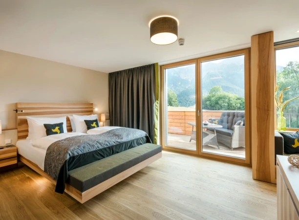 Modern hotel room with a large bed, wooden accents, and a balcony offering mountain views. Cozy outdoor seating enhances the serene atmosphere.