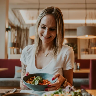 A woman smiling while holding a bowl of food in a cozy restaurant setting.