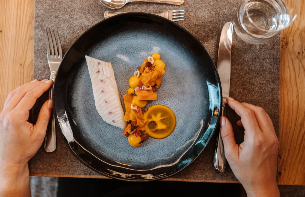Ein Teller mit zartem Fischfilet, buntem Gemüse und einer Zitronenscheibe. Hände bereiten sich zum Essen vor.