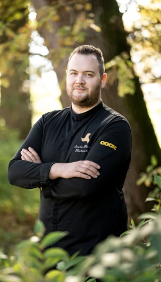 Ein Koch in schwarzer Uniform steht mit verschränkten Armen im Freien vor einem Baum.