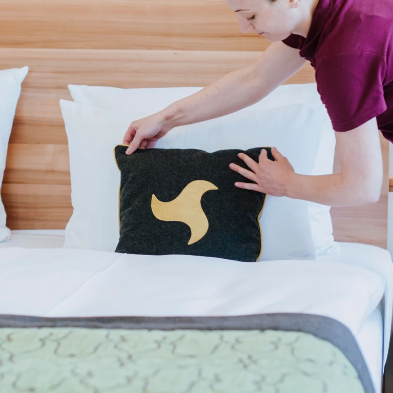Eine Person richtet ein Kissen mit goldenem Design auf einem Bett im Hotel Klosterhof, einem Wellnesshotel in Bayern.