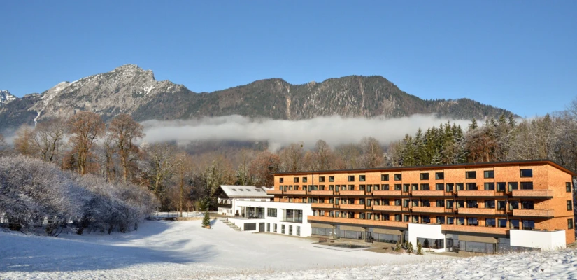 Ein modernes Hotelgebäude umgeben von verschneiten Bergen und Bäumen, unter blauen Himmel und sanften Wolken.