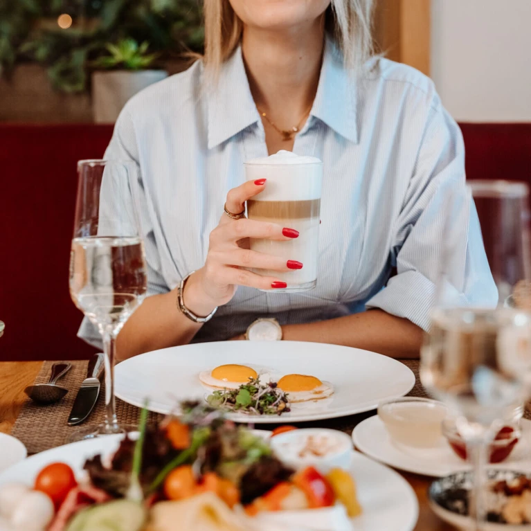 Eine lächelnde Frau mit einem Kaffee in der Hand, umgeben von einem reichhaltigen Frühstückstisch in einem eleganten Restaurant.