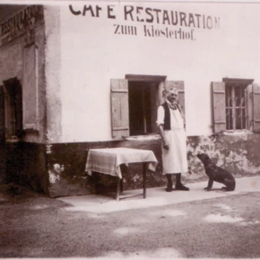 Ein Mann in Schürze steht vor dem Café Restauration zum Klosterhof, neben einem Hund und einem Tisch. Historische Szenerie in Bayern.