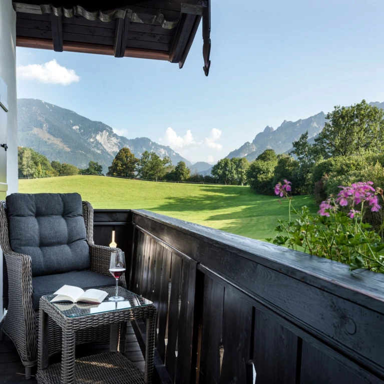 Gemütlicher Balkon der Zirben-Suite im Hotel Klosterhof mit Blick auf die Alpen, einem Sessel, einem Glas Wein und einer Buchseite. Ideal für Entspannung.