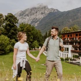 Ein Paar hält Händchen und spaziert durch eine blühende Wiese mit Bergen im Hintergrund, umgeben von Natur und frischer Luft.