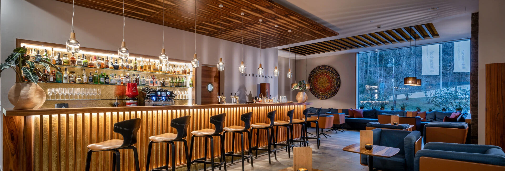 Moderne Bar mit beleuchtetem Tresen, stilvollen Stühlen und gemütlichem Sitzbereich. Fenster mit Blick ins Freie.