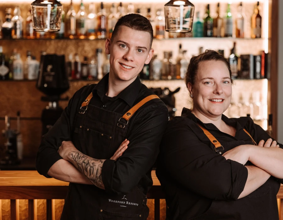 Zwei Barkeeper stehen lächelnd hinter der Bar, umgeben von Spirituosenflaschen. Sie tragen schwarze Kleidung mit orangefarbenen Trägern.