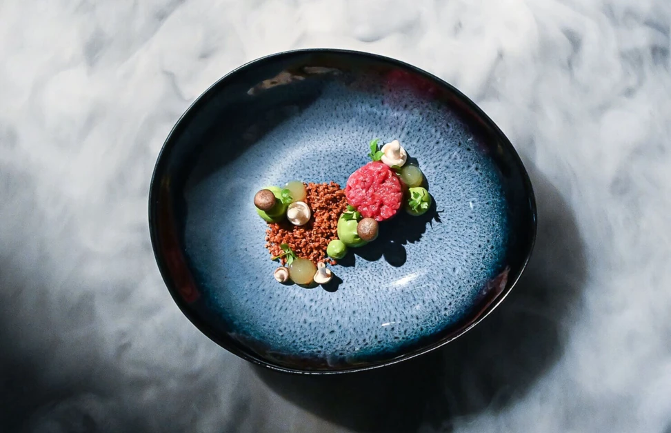 A dish with artistic presentation on a blue plate surrounded by smoke.