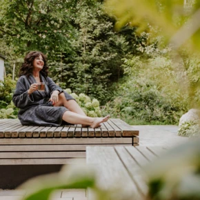 Frau in einem Bademantel sitzt entspannt auf einer Holzliege und genie&szlig;t eine Tasse Tee in einer gr&uuml;nen Gartenlandschaft.