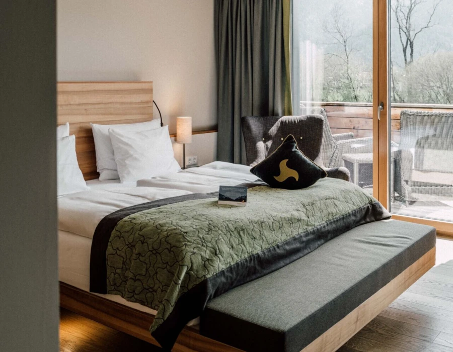 Modernes Hotelzimmer mit großem Bett, eleganter Bettwäsche und Balkon mit Bergblick. Helle Holzakzente und stilvolle Einrichtung schaffen eine gemütliche Atmosphäre.