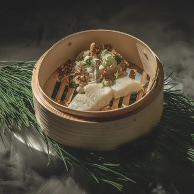 Steamed dish in a bamboo basket garnished with herbs, presented on pine needles, reflecting Hotel Klosterhof's gourmet dining experience.