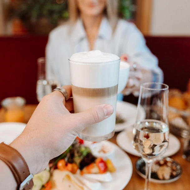 Ein Hand hält ein Glas mit Schaumkaffee, während im Hintergrund eine Frau und ein gedeckter Tisch mit frischem Essen zu sehen sind.