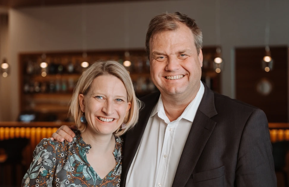 A smiling couple stands together in a warmly lit room with shelves and lights in the background.