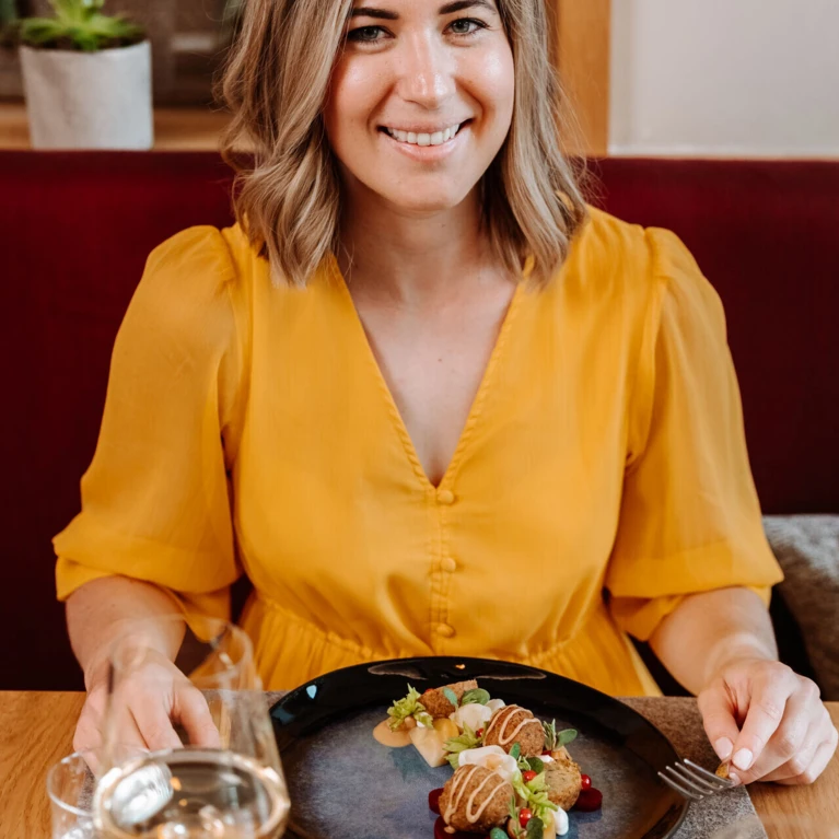 A woman in a yellow blouse sits at a table with a gourmet dish, smiling, in a cozy restaurant setting.
