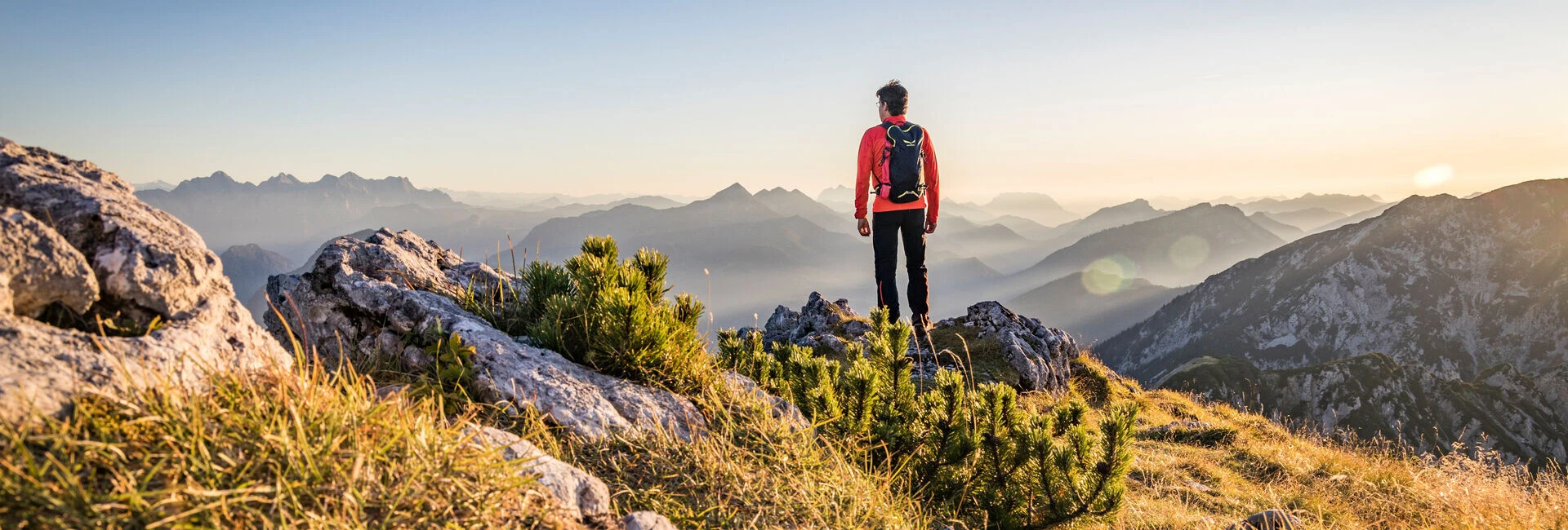 A hiker stands on a grassy mountain peak, overlooking a vast landscape of distant mountains under a clear sky.