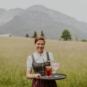 Eine lächelnde Frau in traditioneller Tracht hält ein Tablett mit Getränken vor einer malerischen Berglandschaft.