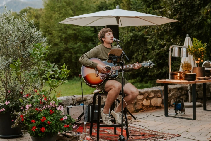 Ein Musiker spielt akustische Gitarre unter einem Sonnenschirm, umgeben von Pflanzen, im Hotel Klosterhof, Bayern.