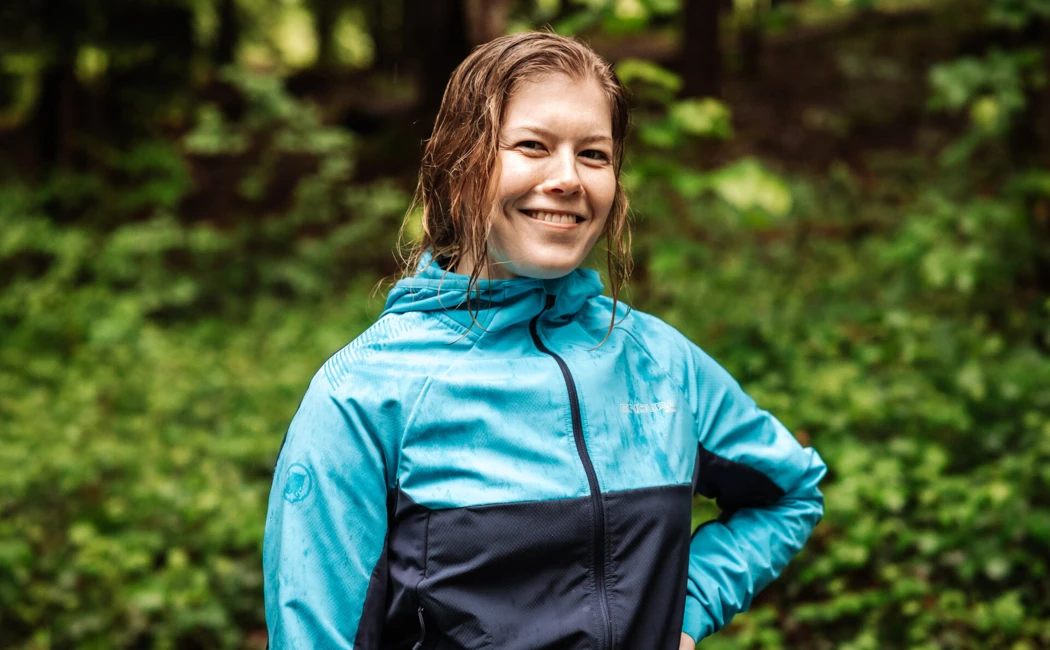 Eine lächelnde Frau in einem blauen Outdoor-Jacket steht im Wald, umgeben von üppigem Grün, ideal für Abenteuer und Erholung.