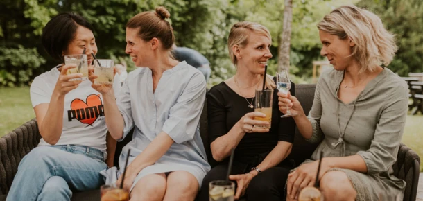 Fünf Frauen genießen Drinks auf einer Couch im Freien, umgeben von grünem Ambiente. Freundliche Stimmung und geselliges Beisammensein.