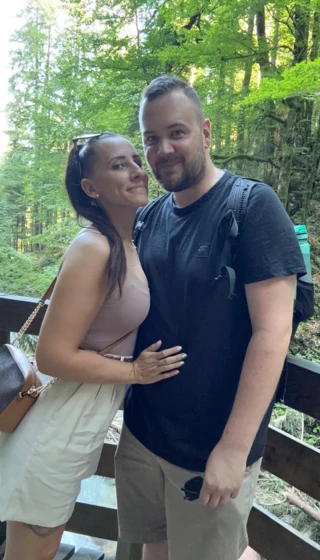 Paar posiert auf einer Holzbrücke im Wald, umgeben von grünen Bäumen. Sie lächeln glücklich in die Kamera.