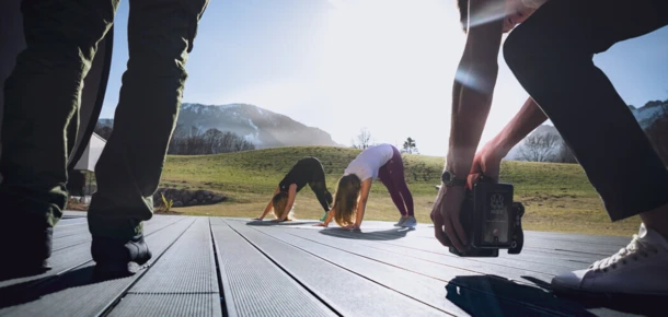 Zwei Frauen machen Yoga auf einer Terrasse im Freien mit Bergen im Hintergrund, während eine Person fotografiert. Hotel Klosterhof, Bayern.
