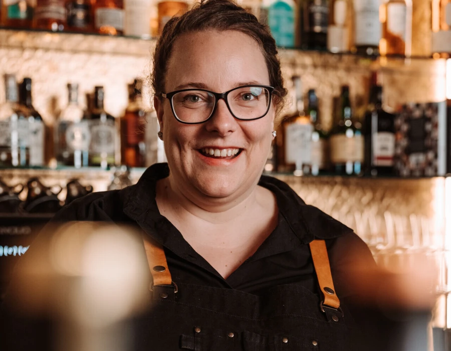 Lächelnde Barkeeperin im Hotel Klosterhof, umgeben von Spirituosen, strahlt Gastfreundschaft und alpine Atmosphäre aus.