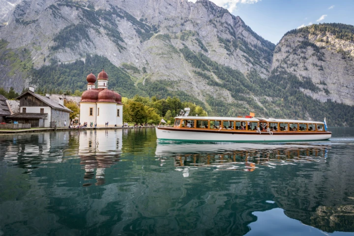 Ein Boot fährt am klaren Wasser vorbei, mit Blick auf die malerische Kirche und die umliegenden Berge im Klosterhof.