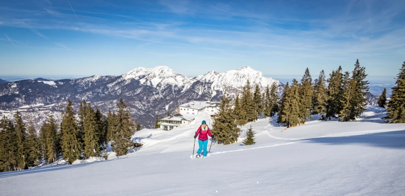 A person snowshoeing on a snowy mountain with evergreen trees and distant peaks, near Wellnesshotel Klosterhof in Bayern.