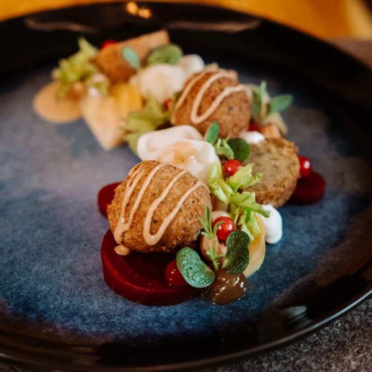A gourmet dish with falafel, beet slices, and fresh greens, elegantly plated with creamy sauce drizzles on a dark plate.