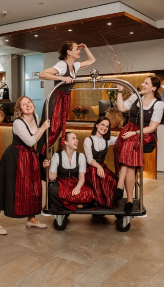 Fünf Frauen in Trachten stehen lächelnd auf einem Gepäckwagen im Hotel Klosterhof, umgeben von einem einladenden Ambiente.