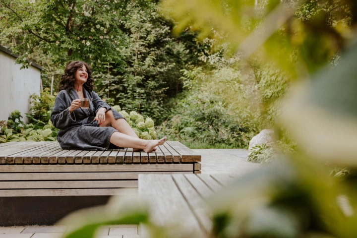 Woman in a robe enjoying tea on a wooden deck in a lush garden setting.