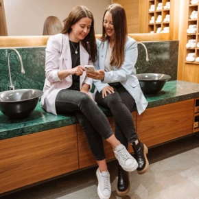 Zwei Frauen in modernen Blazern sitzen am Waschbecken, lächelnd auf ein Handy schauend, in einer stilvollen Wellnessumgebung im Hotel Klosterhof.