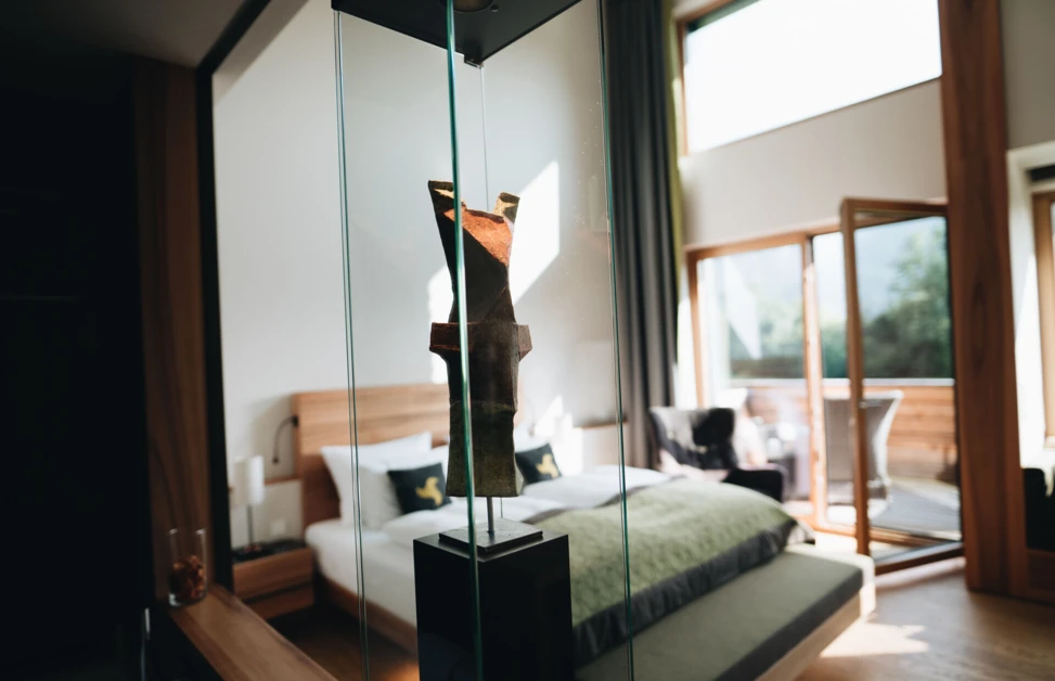 Modern hotel room with a sculpture in the foreground, featuring a cozy bed and open balcony doors leading to a scenic view at Hotel Klosterhof.