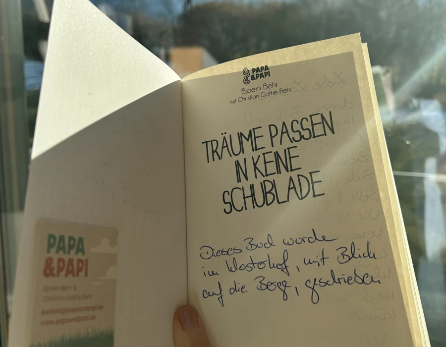 Ein offenes Buch mit dem Titel „Träume passen in keine Schublade“ vor einem Bergpanorama. Handgeschriebene Notiz auf der Innenseite.