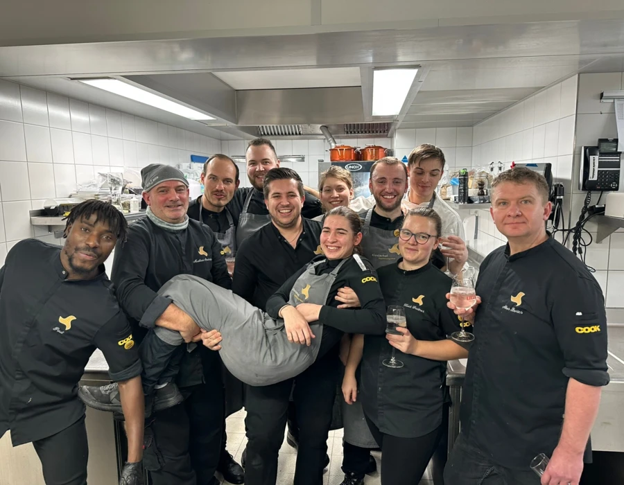 Ein fröhliches Küchenteam im Hotel Klosterhof, umgeben von Kochutensilien, feiert gemeinsam in einer modernen Küche.