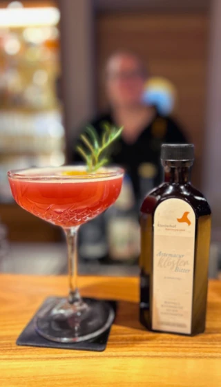 Cocktail mit Rosmarin-Garnitur neben einer Flasche Klosterhof Bitter auf einer Holztheke.