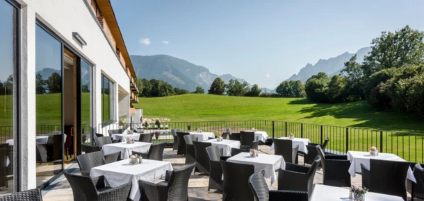 Gemütliche Terrasse mit Tischen und Stühlen, umgeben von grüner Wiese und Bergen im Hintergrund. Ideal für entspannte Mahlzeiten im Freien.
