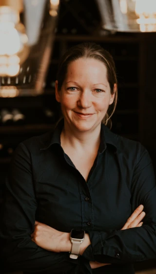 Porträt einer lächelnden Frau mit verschränkten Armen, im Hintergrund eine stilvolle Weinkeller-Atmosphäre.