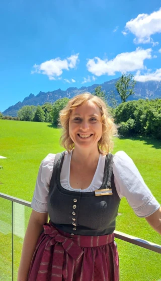 Eine lächelnde Frau trägt ein traditionelles bayerisches Dirndl vor einer malerischen Landschaft mit Bergen und grünem Gras.