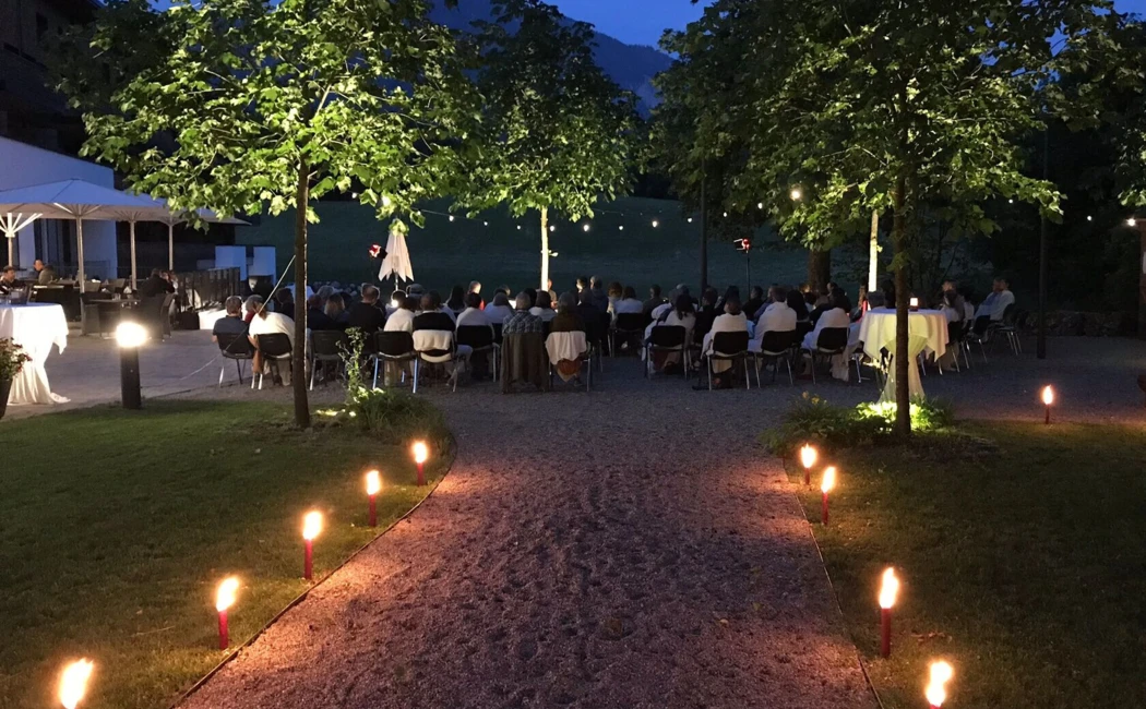 Eine stimmungsvolle Veranstaltung im Freien mit G&auml;sten, umgeben von B&auml;umen und Fackeln, bei D&auml;mmerlicht.