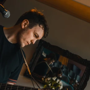 Ein Musiker spielt Gitarre in einer stilvollen Umgebung, mit einem Spiegel im Hintergrund, der die Atmosphäre des Raumes reflektiert.