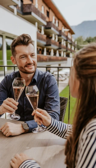 Ein Paar st&ouml;&szlig;t mit Sektgl&auml;sern an, w&auml;hrend sie auf der Terrasse des Hotel Klosterhof sitzen, umgeben von der sch&ouml;nen Alpenlandschaft.