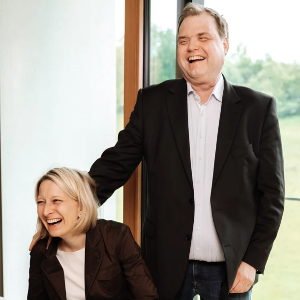 Lachendes Paar in ansprechender Umgebung, entspannt vor großen Fenstern mit Blick ins Grüne.